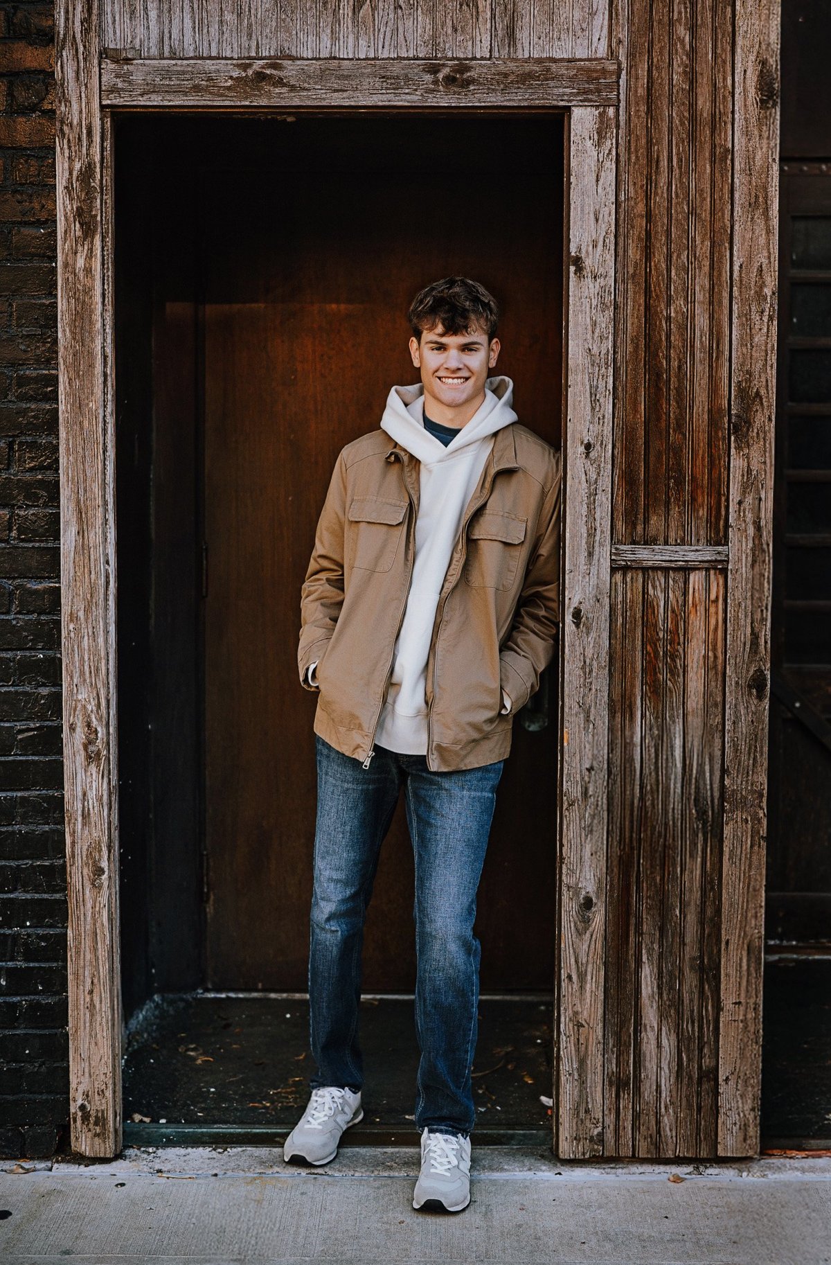 Young man smiling in doorway wearing tan jacket, white hoodie, jeans, and white sneakers against weathered wooden door